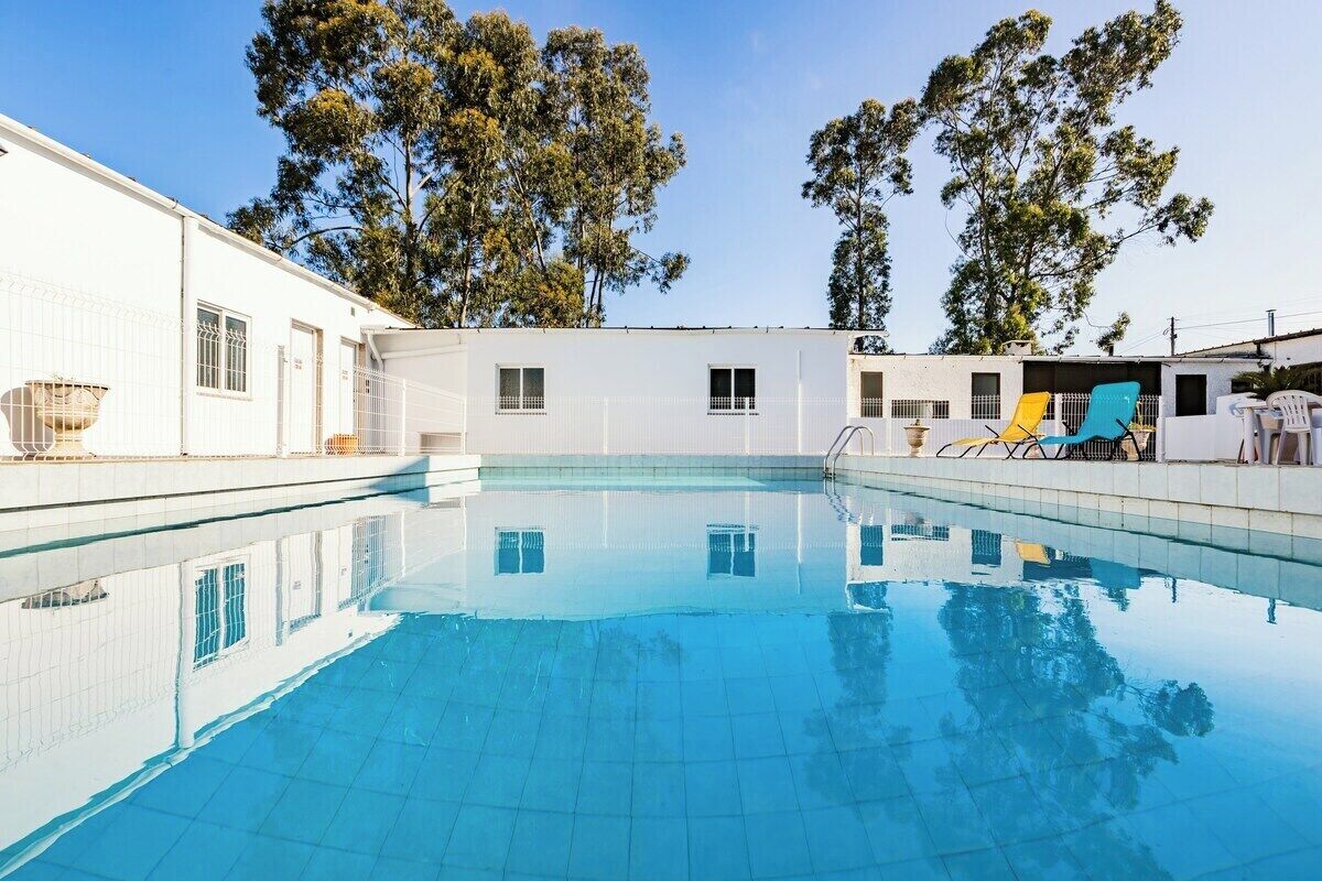 A swimming pool with clear blue water, surrounded by a white building with windows and doors, and trees in the background.