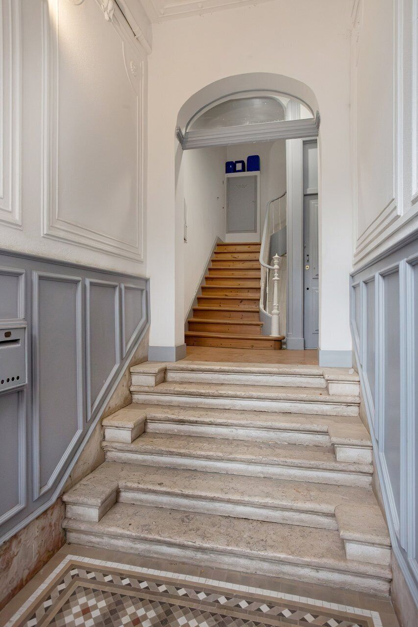 Uma escada leva a uma porta com uma placa azul, com corrimão e piso estampado.