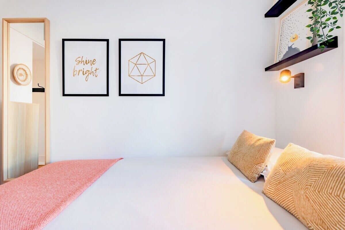 Une chambre à coucher comprenant un lit, des oreillers, une table de chevet, une étagère fixée au mur avec une plante, et deux tableaux encadrés.