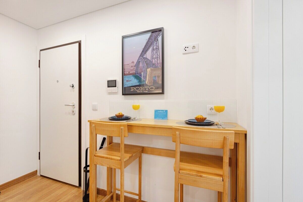 Una habitación con un escritorio de madera, dos sillas, un cuadro enmarcado de un puente, una puerta y una pequeña mesa con un plato y un vaso de jugo de naranja.