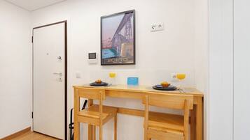 Una habitación con un escritorio de madera, dos sillas, un cuadro enmarcado de un puente, una puerta y una pequeña mesa con un plato y un vaso de jugo de naranja.