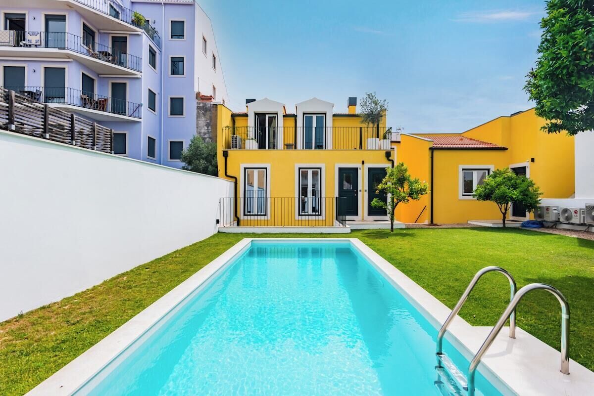 A swimming pool with a white deck and metal railings, surrounded by a well-maintained lawn and a building with a yellow facade and balconies.