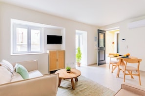 A modern living room with a sofa, a round wooden table with a plant, a television mounted on the wall, and a dining area with chairs and a table. - Lapa Getaway Studio with Swimming Pool (Lisbon)