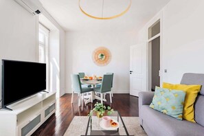 A living room with a flat-screen TV, a dining table with chairs, a sofa with cushions, and a decorative wall piece. - Saldanha Charm Hideaway (Lisbon)
