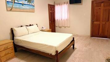 Une chambre à coucher avec un lit, des tables de chevet en bois, une commode, un tableau représentant une scène de plage, un téléviseur fixé au mur et une porte avec un rideau.