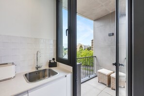 A modern kitchen with a stainless steel sink, a toaster, and a window leading to a balcony with wicker furniture. - Sage 1BR | Balconies, AC & Parking (Porto)