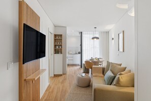 A modern living room with a flat-screen TV, a dining area with a round table, and a kitchen in the background. - Sage 1BR | Balconies, AC & Parking (Porto)