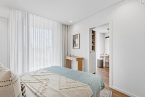 A bedroom with a bed, a bedside table, a desk, a chair, a wardrobe, and a bathroom visible through an open door. - Sage 1BR | Balconies, AC & Parking (Porto)