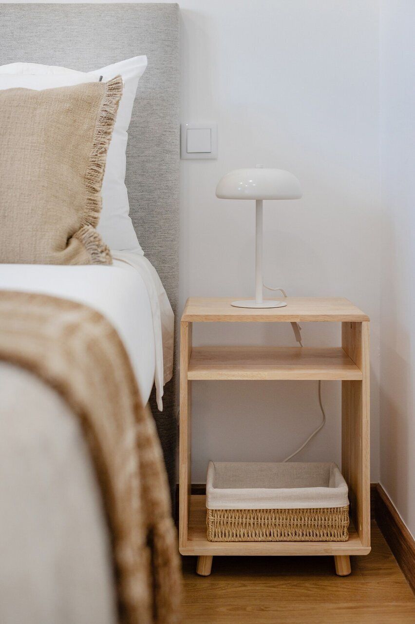 Une chambre à coucher avec un lit, une lampe de chevet, une table de chevet avec un panier, et une prise électrique au mur.