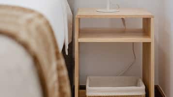 Un dormitorio con cama, lámpara de noche, mesita de noche con una cesta y un enchufe en la pared.