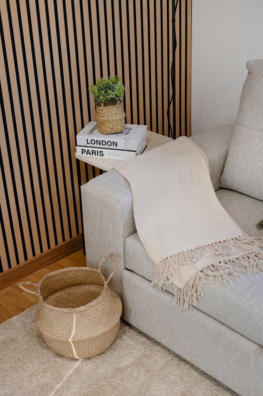 Una sala de estar con un sofá, una cesta tejida, una pequeña mesa con una planta y una pila de libros titulados 'LONDON PARIS'.