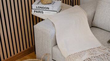 Una sala de estar con un sofá, una cesta tejida, una pequeña mesa con una planta y una pila de libros titulados 'LONDON PARIS'.