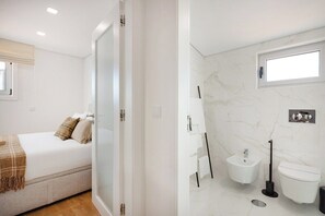 Een slaapkamer met een bed, nachtkastje en raam. De badkamer heeft een toilet, een bidet, een wastafel en een spiegel.