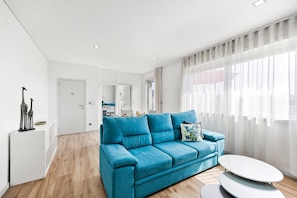 Ein modernes Wohnzimmer mit einem türkisfarbenen Sofa, einem runden weißen Couchtisch und einem großen Fenster mit transparenten Vorhängen.