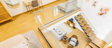 Un interior moderno con un puente de cristal que une dos niveles, un sofá, una mesa con sillas y una escalera.