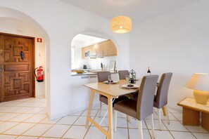 Un coin repas avec une table en bois, six chaises, un luminaire suspendu et une cuisine visible à travers une arche.