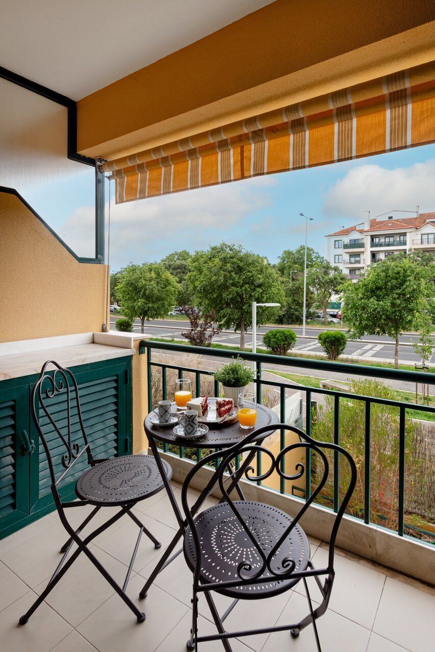 A balcony with two chairs and a small table, a window with a striped blind, and a view of a street and buildings.