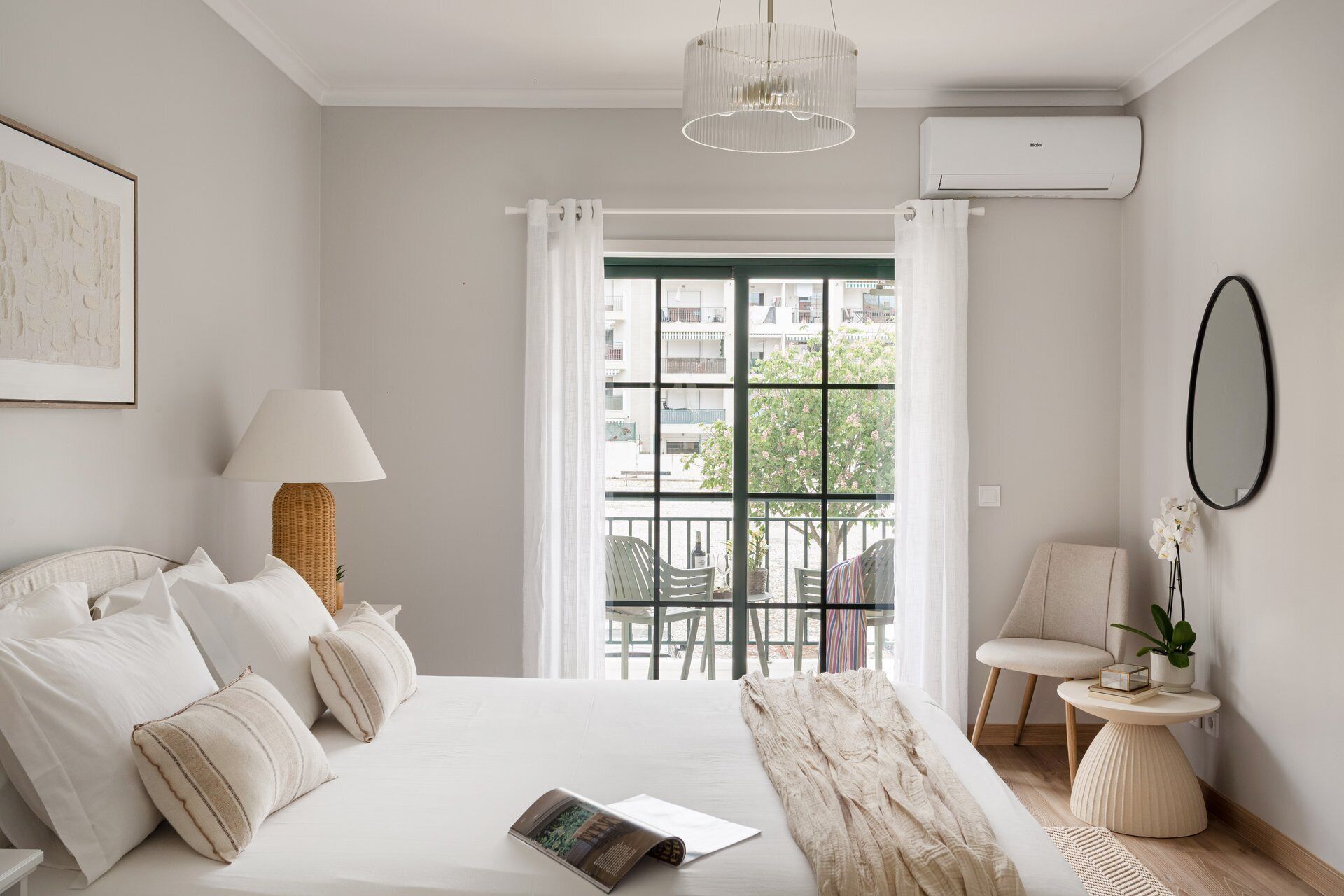 A bedroom with a bed, a window with a view, a chair, a table, and an air conditioning unit.