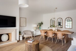 A living room with a fireplace, a dining area with wicker chairs, a round coffee table, and a TV mounted on the wall. - Carcavelos Terrace & Beach Apartment (Carcavelos)