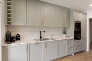 Eine Küche mit hellgrauen Schränken, einer Edelstahl-Spüle, einem schwarzen Wasserhahn, einem eingebauten Backofen und einer Arbeitsplatte mit einem Toaster und einer kleinen Pflanze.