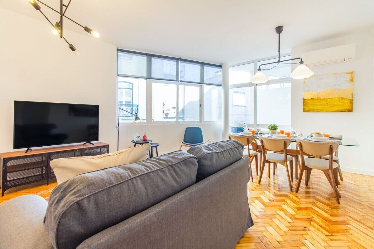 A modern living room with a large flat-screen TV, a sofa, dining area with chairs, and large windows.