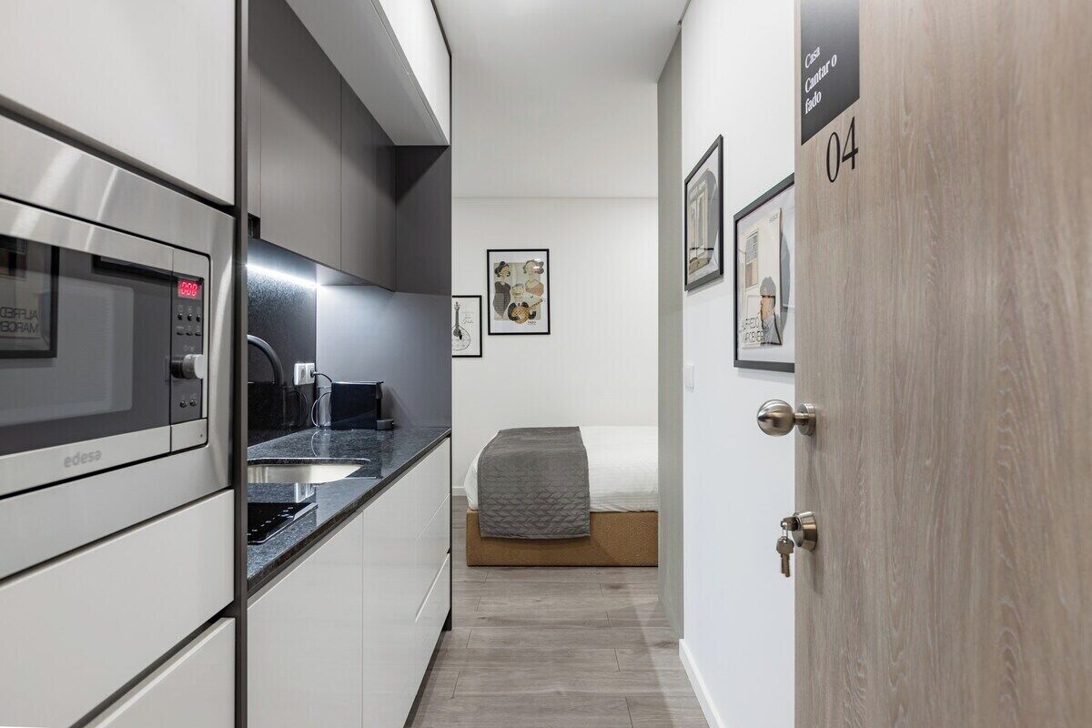 A compact kitchen with a microwave, a coffee machine, and a sink. A bed with a gray blanket is visible in the background.