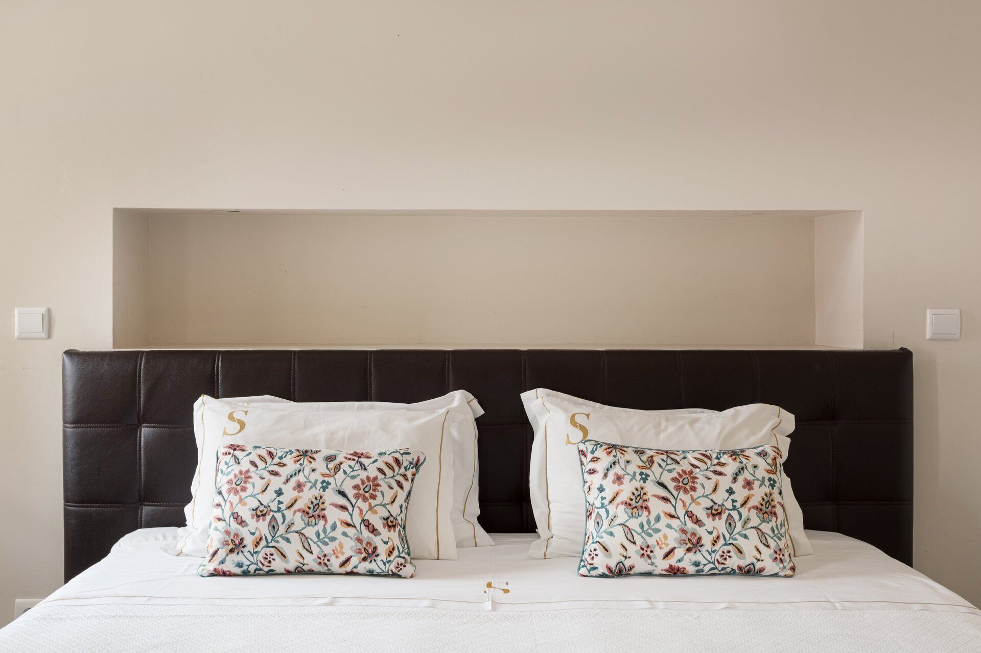 Ein Bett mit dunklem Kopfteil, zwei dekorative Kissen mit floralem Muster und einer schlichten Wand.