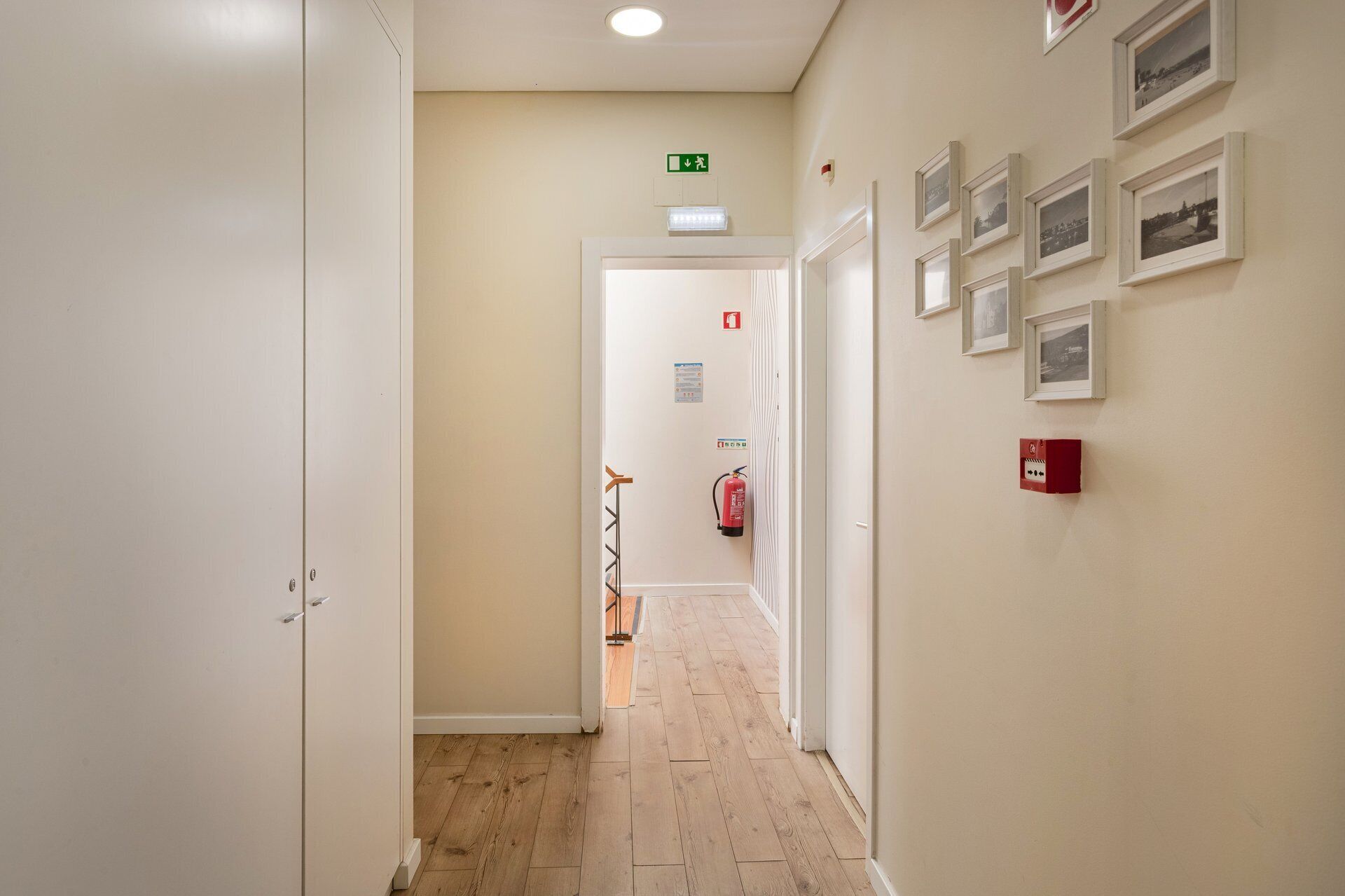 Un pasillo con piso de madera, paredes blancas y cuadros enmarcados a la derecha. Se observa un extintor y un cartel en la pared.