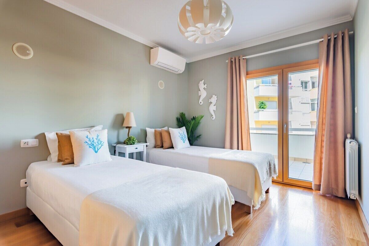 A hotel room with two beds, a ceiling fan, an air conditioning unit, a small table with a lamp, and a view of the outside balcony.