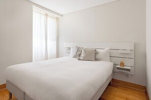 Una cama bien hecha con cabecera blanca, una mesita de noche con lámpara y una ventana con cortinas.