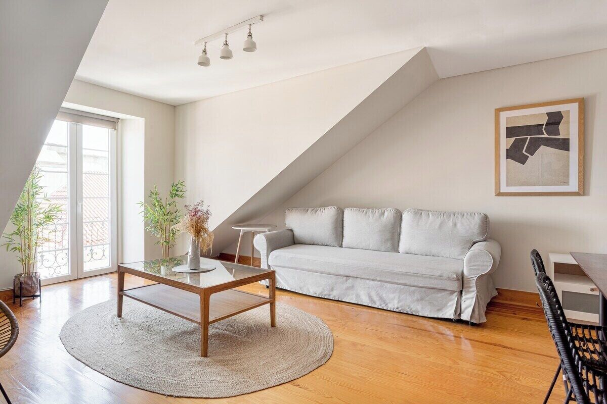 A modern living room with a white sofa, a glass coffee table, and a dining table with chairs. There is a large window providing natural light and a view of the outside.