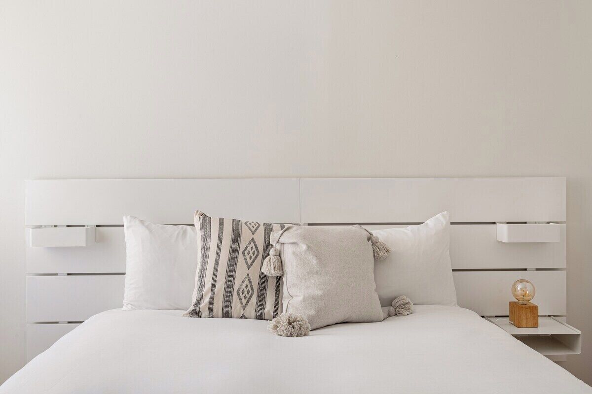 A bed with white bedding, a patterned pillow, and a small wooden nightstand with a glass object.