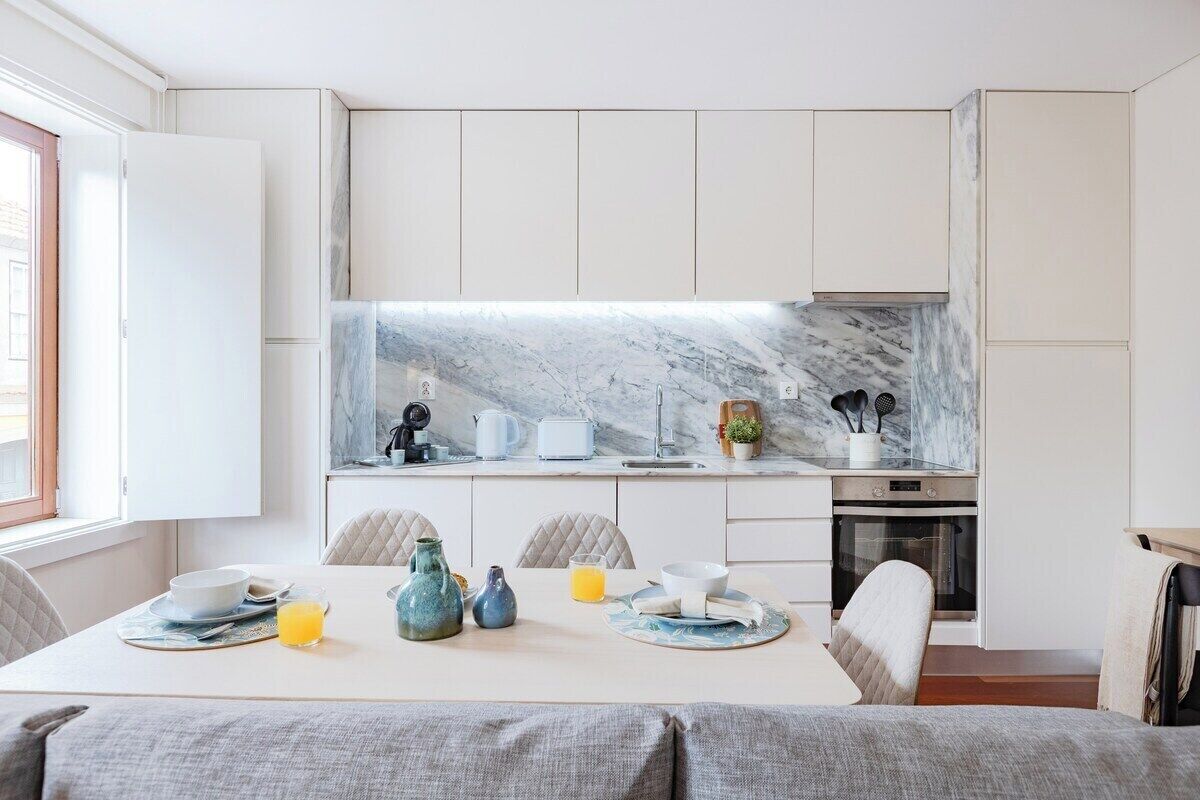 Une cuisine moderne avec des meubles blancs, un pare-feu en marbre et un coin repas où une table est dressée pour le petit-déjeuner.
