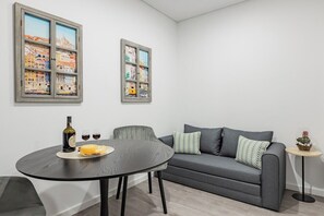 A modern living room with a grey sofa, a round table with a bottle and glasses, and two framed cityscape paintings on the wall. - Arte do Azulejo @ Boavista Executive Studios (Porto)