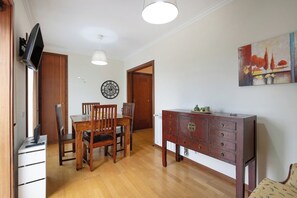 Een eetgedeelte met een houten tafel en stoelen, een wit kastje met lades, een klok aan de muur en een schilderij.