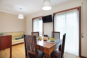 A dining area with a wooden table, four chairs, a television mounted on the wall, and a sofa in the background. - Espinho Retreat with Balcony (Espinho)