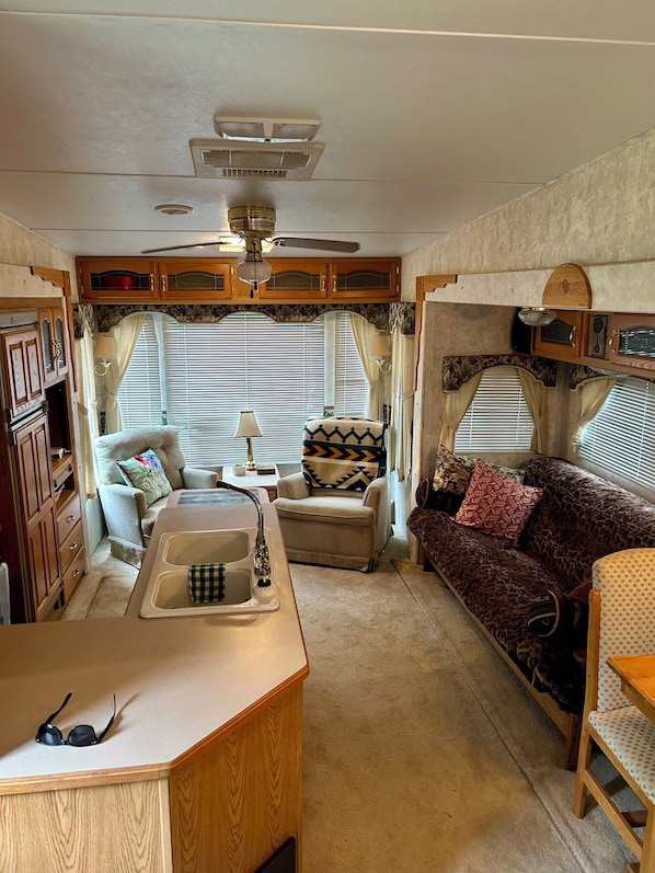 Interior compacto de una caravana con kitchenette, ventilador de techo, sofá y ventana con persianas.