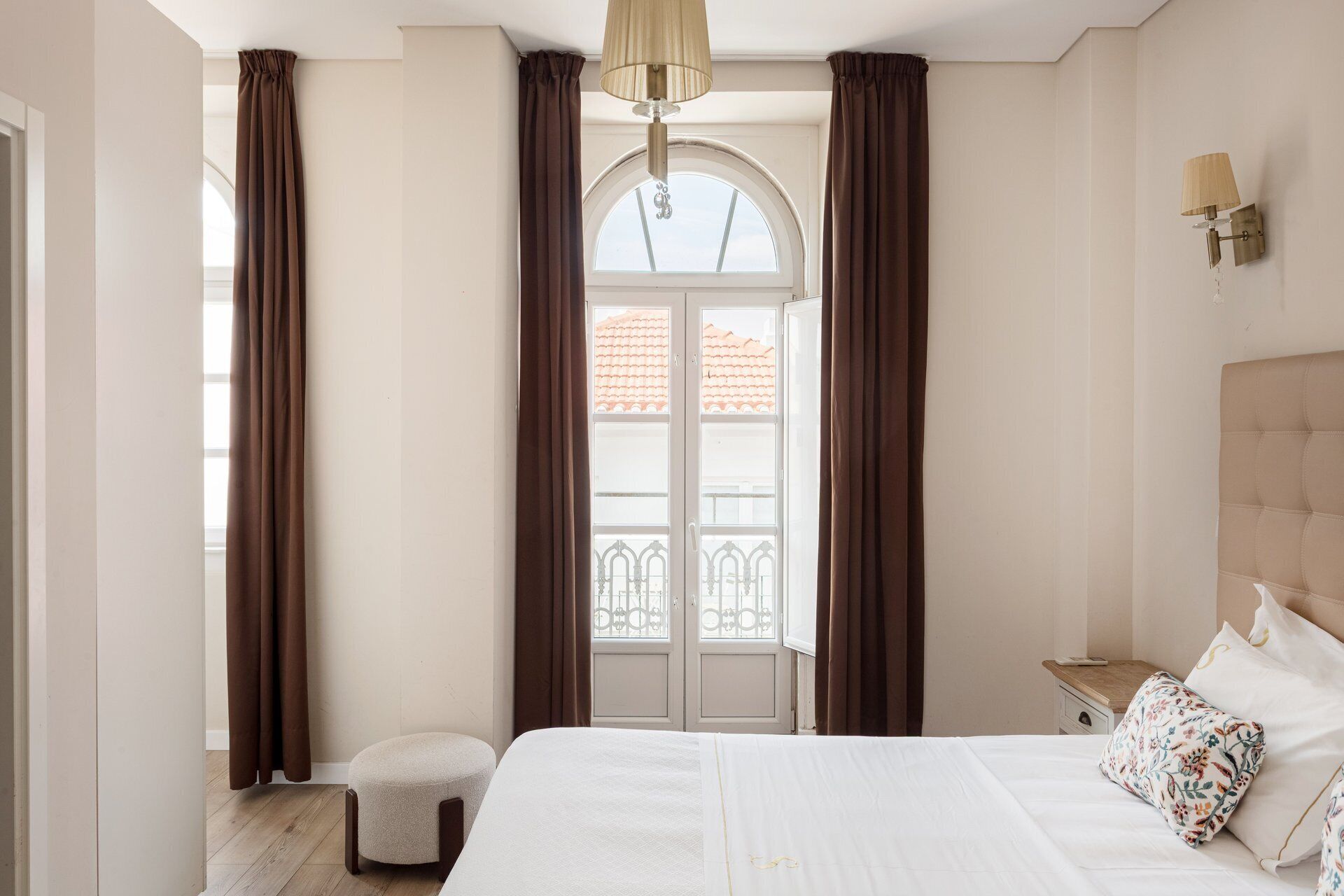 A bedroom with a bed, a nightstand, a lamp, a window with curtains, and a balcony view.