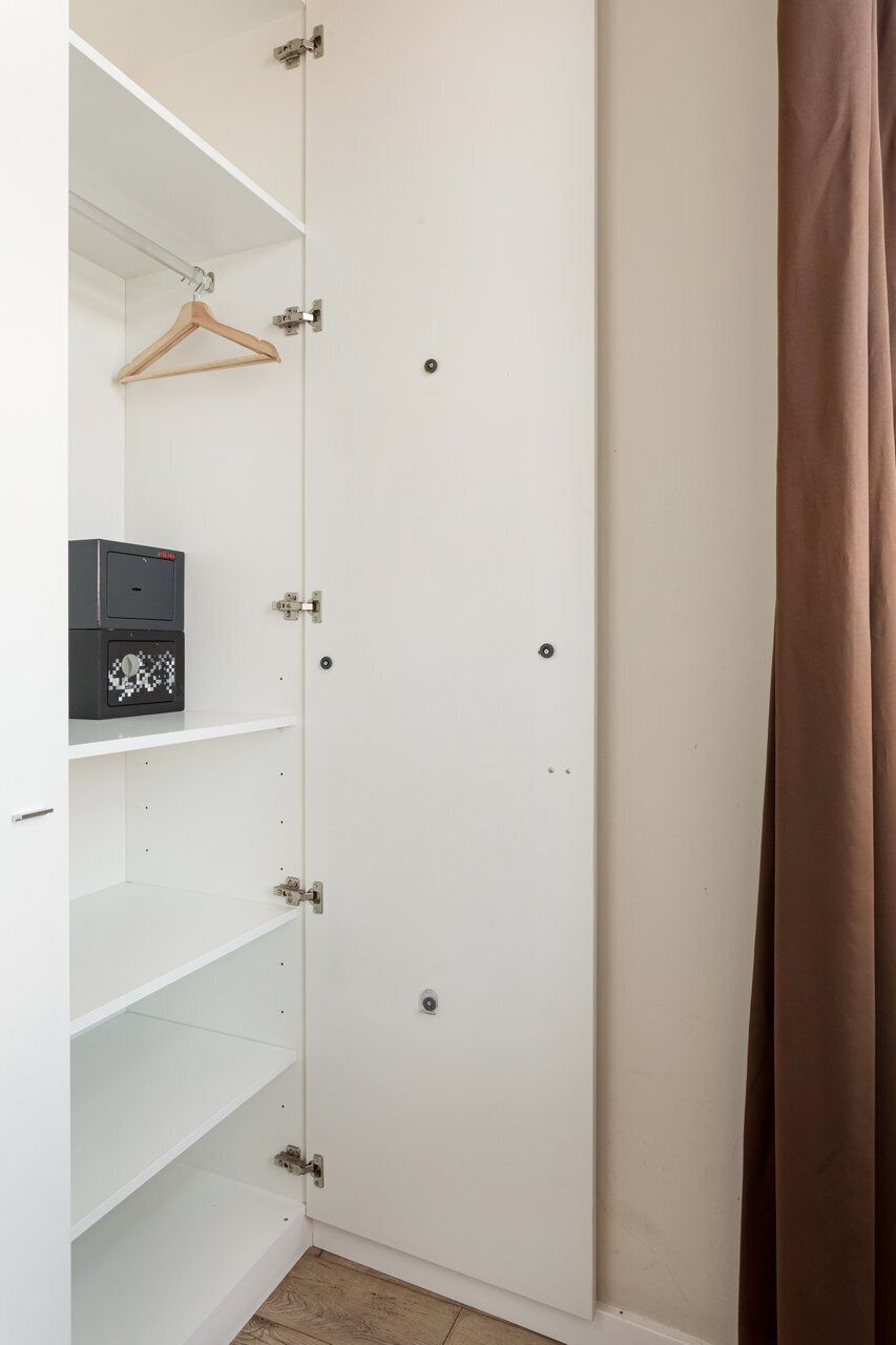 Ein weißer Kleiderschrank mit mehreren Regalen und einer Kleiderstange, eine schwarze Schachtel auf dem obersten Regal sowie ein brauner Vorhang, der teilweise zu sehen ist.