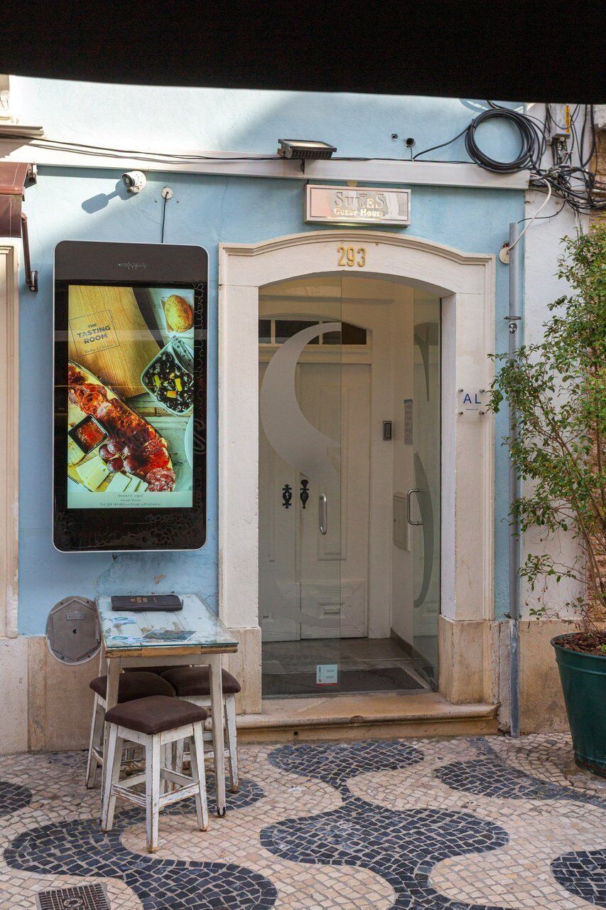 A guest house with a glass door entrance, a sign above the door, a small table with a chair, and a potted plant to the right.