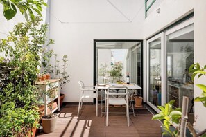 Een terras buiten met een tafel en stoelen, omgeven door potplanten en een uitzicht door een glazen deur.