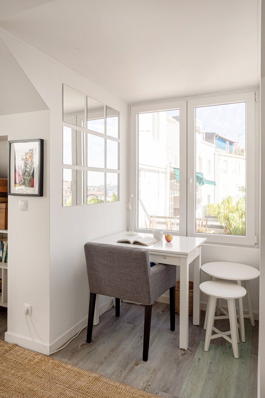 Una habitación con una mesa blanca, una silla gris, un taburete pequeño y un ventanal con vistas a edificios y vegetación.