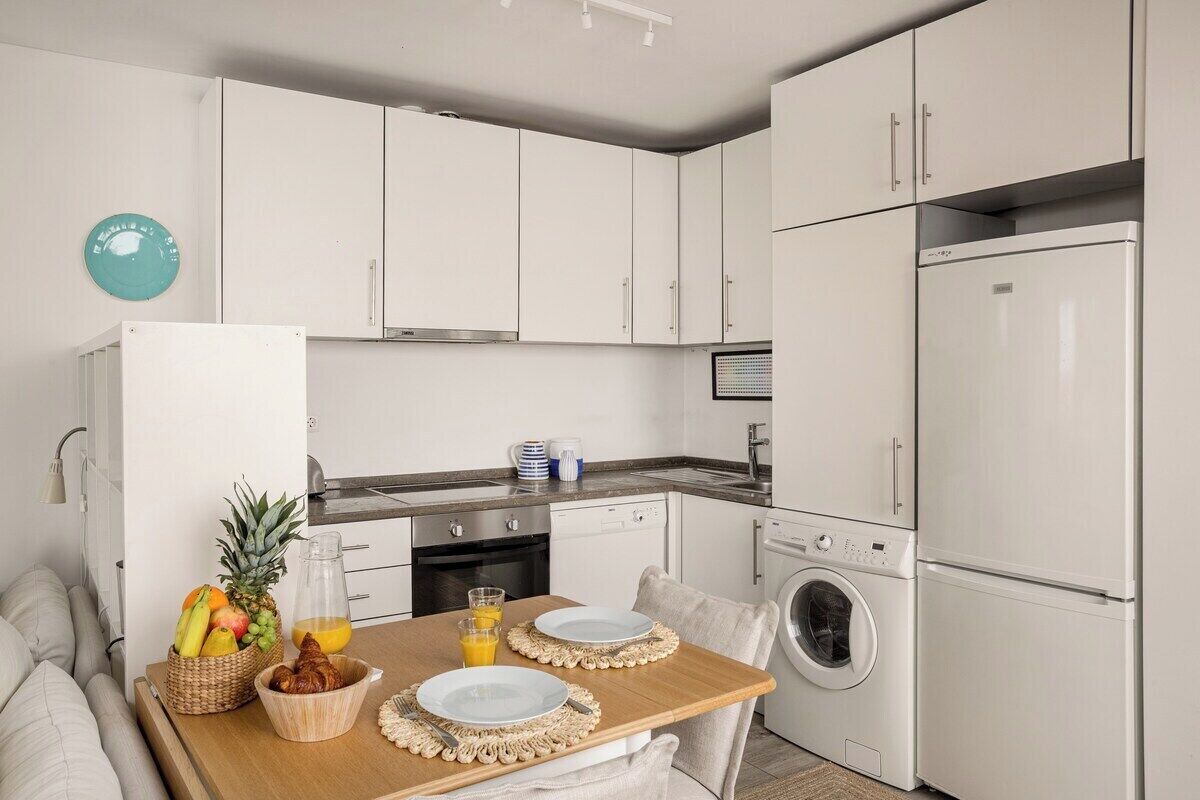 A modern kitchen with white cabinets, a built-in oven, a washing machine, and a dining table set with plates, a fruit bowl, and a juice pitcher.