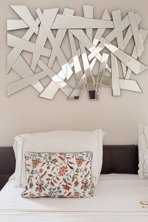 Un lit avec une literie blanche, un coussin à motifs floraux et une œuvre d’art abstraite en forme de miroir mural.