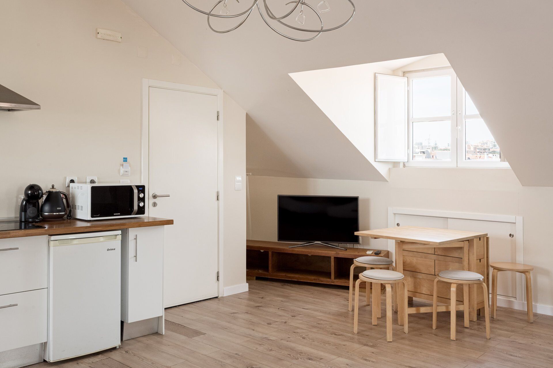 Eine kompakte Küche mit Mikrowelle, Kaffeemaschine und kleinem Kühlschrank. Ein Fernseher steht auf einem Holzboden und es gibt einen Essbereich mit Hocker.