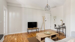 Een moderne woonkamer met een flatscreen televisie, een houten salontafel, een grijze bank en een eethoek met een klein tafeltje en stoelen.