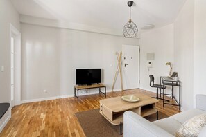 Una sala moderna con un televisor de pantalla plana, una mesa de centro de madera, un sofá gris y un comedor con una mesa pequeña y sillas.
