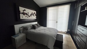 Un dormitorio con cama, mesita de noche, banco, armario y una obra de arte en la pared.