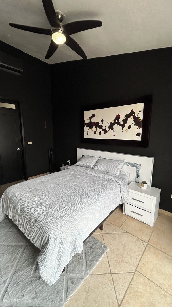 Une chambre à coucher comprenant un lit, des tables de chevet, un ventilateur de plafond, une œuvre d’art accrochée au mur et un sol carrelé.