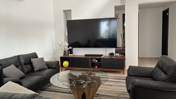 Una sala de estar moderna con un televisor de pantalla plana grande, dos sofás de cuero negro, una mesa de centro de vidrio y un mueble de entretenimiento de madera.